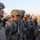 A group of U.S. Army soldiers in ACU (Army Combat Uniform) digital camouflage are standing in formation, appearing to be at attention or receiving a briefing. The soldiers are fully equipped with helmets, body armor, and tactical gear. The setting appears to be an outdoor military environment, likely a base or staging area, during early morning or late afternoon light.