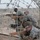 The image shows multiple military personnel in camouflage uniforms working in a sandy, arid environment behind concertina/razor wire. The soldiers appear to be engaged in construction or fortification work, handling wooden planks or boards. The scene is photographed through the razor wire, which partially obscures the view.