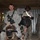 A group of military personnel in US Army ACU (Army Combat Uniform) camouflage are gathered in what appears to be an indoor military facility or common area. The central figure is carrying a rifle and holding what appears to be a saddle, suggesting a themed event or ceremony. A banner in the background partially reads 'CSM Some... Cigar Ni...' indicating this may be a Command Sergeant Major recognition or social event.