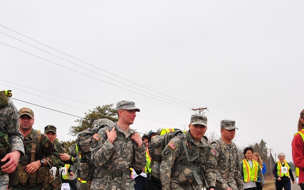 A group of U.S. Army soldiers in ACU (Army Combat Uniform) carrying rucksacks are participating in what appears to be a ruck march or charity walk/run event, identifiable by numbered race bibs attached to their uniforms. Civilian participants wearing high-visibility safety vests and casual athletic wear are also present alongside the soldiers on a paved road. The overcast sky and bare trees suggest a cool-weather setting, likely early spring or late fall.