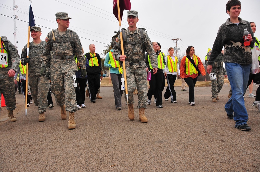 A group of people are participating in an organized walk or ruck march event on a paved road. Two soldiers in US Army ACU (Army Combat Uniform) camouflage are leading the group, one carrying a maroon/burgundy guidon flag and both wearing rucksacks. Civilian participants wearing numbered bibs and high-visibility safety vests follow behind them.