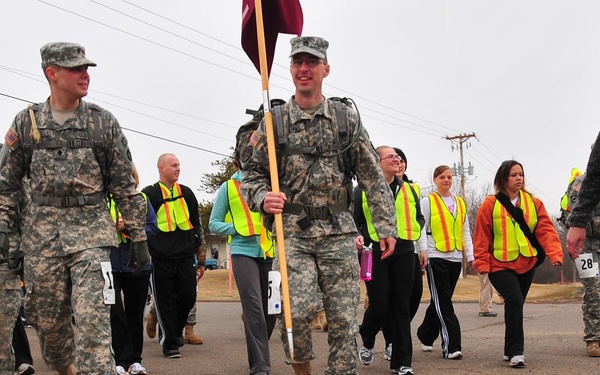 A group of people are participating in an organized walk or ruck march event on a paved road. Two soldiers in US Army ACU (Army Combat Uniform) camouflage are leading the group, one carrying a maroon/burgundy guidon flag and both wearing rucksacks. Civilian participants wearing numbered bibs and high-visibility safety vests follow behind them.