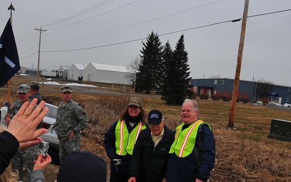 An outdoor gathering at what appears to be a military installation or base, where civilians wearing high-visibility safety vests are posing for photographs alongside military personnel in camouflage uniforms. Several people are taking photos of a small group posing together, including an elderly individual flanked by two people in safety vests. The overcast sky and bare vegetation suggest a cold-weather season, likely late fall or early spring.