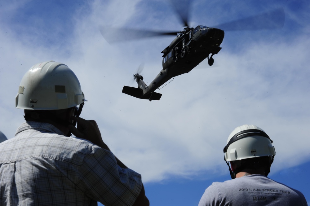 Two individuals wearing white hard hats observe a military helicopter flying at low altitude overhead. The helicopter appears to be a UH-60 Black Hawk, captured mid-flight against a partly cloudy blue sky. One observer appears to be photographing or communicating via radio while watching the aircraft.