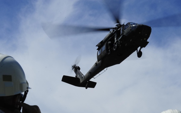 Two individuals wearing white hard hats observe a military helicopter flying at low altitude overhead. The helicopter appears to be a UH-60 Black Hawk, captured mid-flight against a partly cloudy blue sky. One observer appears to be photographing or communicating via radio while watching the aircraft.