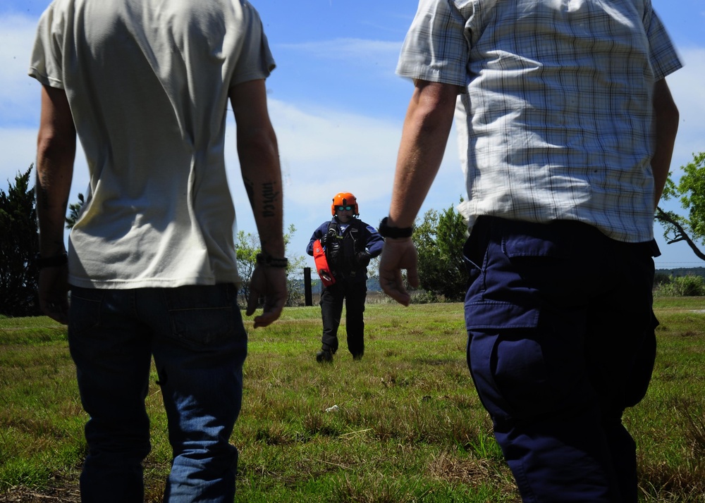 Three individuals are standing in an open grassy field under a partly cloudy sky. The central figure is wearing an orange safety helmet, sunglasses, and a dark tactical vest with equipment, facing two other individuals whose backs are to the camera. The setting and gear suggest a search and rescue training exercise or operational briefing.
