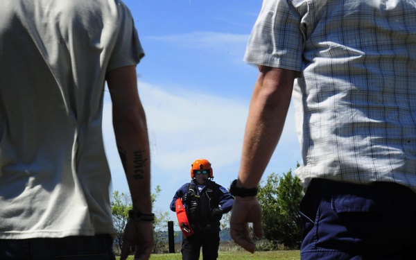 Three individuals are standing in an open grassy field under a partly cloudy sky. The central figure is wearing an orange safety helmet, sunglasses, and a dark tactical vest with equipment, facing two other individuals whose backs are to the camera. The setting and gear suggest a search and rescue training exercise or operational briefing.