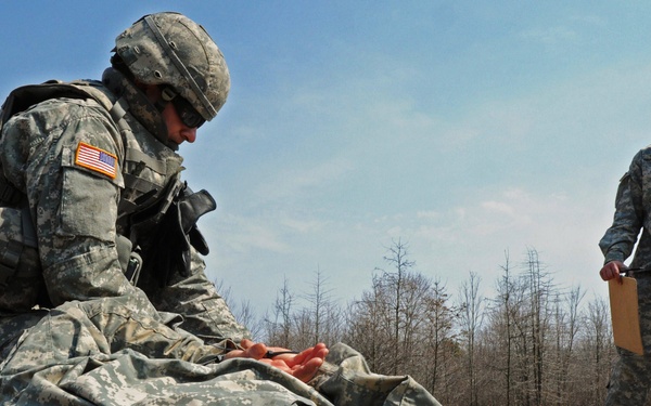Three U.S. Army soldiers are engaged in what appears to be a combat first aid or casualty care training exercise in an outdoor field setting. One soldier in full combat gear is attending to a second soldier lying face-down on a rubber mat, simulating a casualty scenario. A third soldier stands nearby holding a clipboard, likely acting as an evaluator or observer for the training exercise.