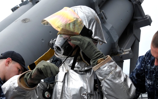Two U.S. Navy sailors in digital camouflage uniforms are assisting a third sailor in donning a silver aluminized proximity firefighting suit aboard what appears to be a naval vessel. The suited individual is adjusting a full-face respirator/gas mask while the others secure straps and equipment. This appears to be a firefighting drill or equipment check on a ship's deck.