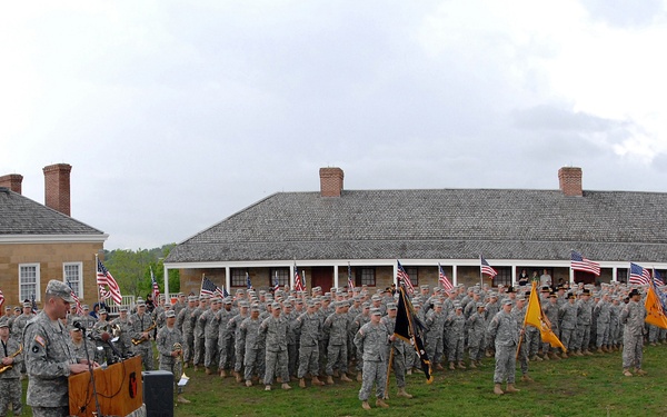 A formal U.S. Army military ceremony is taking place outdoors on a grass parade field, with a large formation of soldiers in ACU (Army Combat Uniform) standing at attention. A uniformed officer is speaking at a podium while color guard units hold unit guidons and American flags. Civilian and military dignitaries are seated in the foreground observing the ceremony, with historic-looking stone/brick barracks buildings visible in the background.