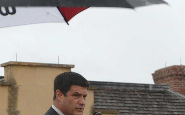 A man in a dark suit and patterned tie is speaking at an outdoor podium equipped with multiple microphones. The wooden lectern displays a red bull skull insignia, suggesting a military or official organizational event. The weather appears overcast and rainy, with umbrellas visible in the background.