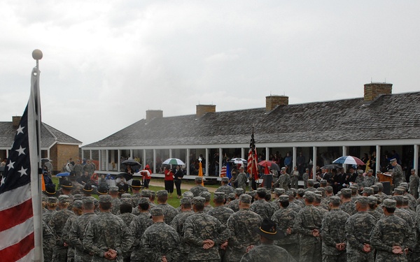 A large military formation ceremony is taking place outdoors at what appears to be a historic fort or military installation on a rainy, overcast day. Numerous soldiers in ACU (Army Combat Uniform) camouflage are assembled in formation facing a raised platform or porch area where officials and dignitaries are gathered. Two American flags are prominently displayed flanking the formation.