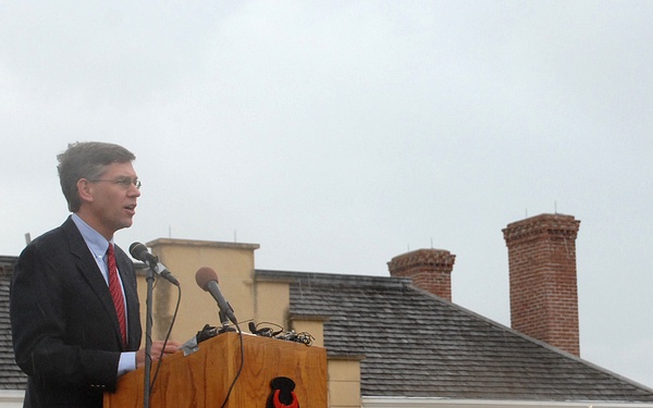 A civilian man in a dark suit and red tie is speaking at a podium during a military ceremony. The wooden podium bears a red bull skull insignia, likely representing the 34th Infantry Division or a similar unit. Multiple uniformed U.S. Army soldiers are assembled in the background, some holding brass instruments, suggesting a military band is present.