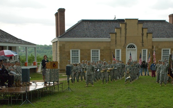 An outdoor military ceremony is taking place on a grass field in front of a large historic stone building, likely a colonial-era structure. A civilian woman is speaking at a podium on a raised platform while military personnel in camouflage uniforms are assembled in formation. The event appears to be occurring during overcast, rainy conditions, with attendees using umbrellas.
