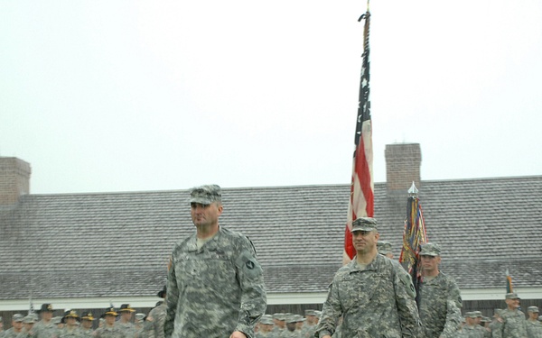 A military formation ceremony is taking place outdoors on a grass field in rainy conditions. Several soldiers in US Army ACU (Army Combat Uniform) camouflage are leading a procession while a large formation of troops stands at attention in the background. An American flag and what appears to be a unit guidon are being carried during what looks like a formal military ceremony.