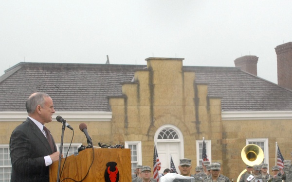 A civilian official in a dark suit is speaking at a podium during an outdoor military ceremony. Behind him stands a formation of U.S. Army soldiers in ACU (Army Combat Uniform) camouflage, several holding musical instruments, indicating a military band is present. The event takes place in front of a historic stone building, with American flags and what appears to be a unit flag visible.