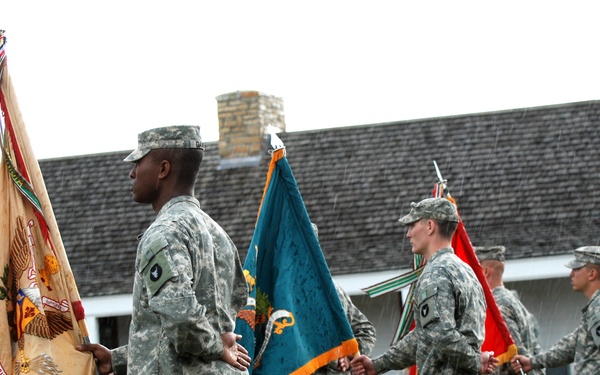 A military color guard ceremony is taking place outdoors on a grass field in rainy conditions. Multiple soldiers in US Army ACU (Army Combat Uniform) camouflage are standing in formation, each holding a different military unit guidon or flag on wooden staffs. The ceremony appears to be a formal military event such as a change of command or unit activation, conducted in front of a historic-looking building.