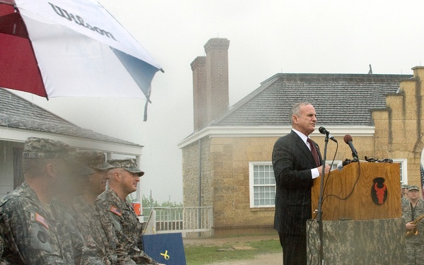 An outdoor military ceremony is taking place in rainy conditions. A civilian man in a dark suit is speaking at a wooden podium bearing a red bull insignia, addressing seated military personnel in camouflage uniforms. The event appears to be a formal military function, likely a change of command, dedication, or commemorative ceremony.