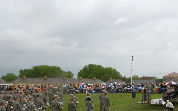 A military ceremony or parade is taking place on a large open parade ground, likely at a historic fort, with soldiers in ACU (Army Combat Uniform) camouflage standing in formation. A military band is visible on the left side with instruments including a bass drum marked '34th Infantry' and brass instruments. Spectators with umbrellas are gathered in the background under overcast, threatening skies.
