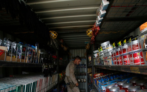 A person in military camouflage uniform is browsing inside what appears to be a small military forward operating base (FOB) store or 'hajji shop' housed in a shipping container. The shelves on both sides are stocked with cigarettes, tobacco products, energy supplements, protein powders, and other consumer goods. The narrow aisle and metal shelving units are characteristic of a makeshift retail space common on military bases.