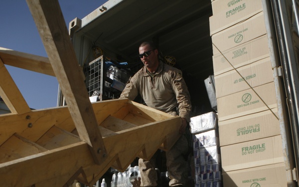Two military personnel in desert camouflage uniforms are unloading supplies from a military vehicle in what appears to be an arid, desert environment. One soldier stands inside the cargo bed while another on the ground handles a wooden pallet or crate structure. The cargo includes boxes marked 'FRAGILE DO NOT DROP' and cases of bottled water.