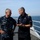 Commanders aboard the weather deck of De Ruyter Two senior naval officers are engaged in conversation on the deck of a naval vessel at sea. One officer wears a US Navy digital camouflage uniform with the name tag 'SHARPE', while the other wears a dark navy uniform with gold rank insignia and what appears to be a Netherlands flag patch. The setting is an open ship deck with calm ocean waters visible in the background.