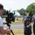 Oceana sailors film local PSA A civilian cameraman is filming two U.S. Navy personnel in Navy Working Uniform (NWU) digital camouflage pattern outdoors. The female sailor is giving a thumbs-up gesture toward the camera while standing alongside a male sailor. A light blue military jet aircraft is visible on display in the background.