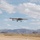 Tiger Shark UAV returns from mission A small fixed-wing unmanned aerial vehicle (UAV) is captured in low-altitude flight over an arid, desert airstrip. The aircraft appears to be either taking off or landing, with landing gear deployed and visible. The background features rugged, barren mountain ranges under a partly cloudy sky.
