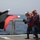 Firefighting exercise aboard USS Stephen W. Groves Naval personnel are conducting what appears to be a firefighting or damage control drill on the deck of a military vessel at sea. Crew members wearing red protective vests, helmets with face shields, and hearing protection are handling a fire hose, while another crew member in the foreground holds signal flags. The scene takes place on an open ship deck with safety netting along the railing and calm ocean visible in the background.