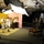 US NF Europe rock band perform on Ukraiane TV A television or music video production is underway in an indoor studio with a colorful set featuring yellow and pink walls. A band dressed in white sailor-style uniforms is performing, with a lead singer at the microphone and musicians playing saxophone and drums. Multiple camera operators and crew members are positioned around the set capturing the performance.