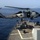 SH-60B Sea Hawk lowers cargo aboard USS Mason A U.S. Navy helicopter, marked 'NAVY' on the fuselage with the number '23' visible, hovers over the flight deck of a naval warship at sea. The helicopter appears to be an SH-60 Seahawk variant performing a deck landing or hover operation. A deck crew member is visible on the ship's flight deck, likely acting as a landing signal officer or safety observer.