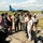 Press conference with Ukraine media A press conference or media interview is taking place on a military airfield tarmac, with journalists holding microphones toward military personnel in flight suits and uniforms. In the background, a blue camouflage fixed-wing aircraft (likely an Antonov An-26) and a blue helicopter (likely a Mil Mi-14 or similar) are visible on the runway. The scene suggests a multinational military event with both local media and international military participants.