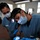 Continuing Promise 2011 Two medical/dental professionals in blue scrubs and surgical masks are performing a dental procedure on a reclined patient. The primary practitioner is wearing an Energizer brand headlamp for illumination and blue nitrile gloves while working in the patient's mouth. The setting appears to be a makeshift or field dental clinic, possibly in a resource-limited environment.