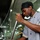 USS Ronald Reagan galley A chef in a gray uniform with gold trim and black buttons is stirring a large metal pot using a long stainless steel rod in a commercial kitchen. A second kitchen worker wearing a white cap is visible in the background. The setting appears to be a large-scale institutional or ship galley kitchen based on the stainless steel equipment and confined space.