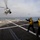HSM 77 USS Freedom joint exercise Two naval personnel in yellow safety vests and helmets are directing a military helicopter approaching a ship's flight deck for landing. The scene takes place on what appears to be a Littoral Combat Ship (LCS), identifiable by the 'LCS' markings on the crew's vests. A second helicopter is visible in the background over open ocean.