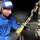 Airman aboard USS Ronald Regan chains a F/A-18C Hornet A technician wearing a blue helmet with hearing protection and a blue sweatshirt is working on what appears to be aircraft landing gear or a tie-down assembly. They are handling a yellow and silver tensioning device connected to a chain and hook mechanism. The setting appears to be inside an aircraft hangar or carrier deck bay.