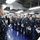 USS Bataan frocking ceremony A senior U.S. Navy officer is addressing a large formation of sailors aboard what appears to be a naval vessel, likely a ship's hangar bay or interior deck space. The officer is holding a microphone and speaking to rows of personnel standing at attention in Navy Working Uniform (NWU) digital camouflage. Several sailors are holding white documents or certificates, suggesting this may be an awards ceremony, promotion ceremony, or official briefing.