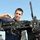 USS San Antonio sailor cleans machine gun A U.S. Navy sailor in blue utility uniform is performing maintenance on a mounted crew-served weapon, likely an M2 .50 caliber machine gun or similar heavy weapon system. The work appears to be conducted on the deck of a naval vessel, with a second sailor visible in the background. The sailor is using a tool, possibly a brush or screwdriver, to clean or adjust a component of the weapon.