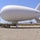 PGSSS aerostat undergoes testing A large white tethered aerostat (blimp) marked '22M.041' is grounded in an arid desert environment, likely a southwestern U.S. location. The aerostat is secured to ground equipment and mooring lines, with safety cones marking the perimeter. A single person stands near the ground support equipment, providing scale reference to the substantial size of the craft.