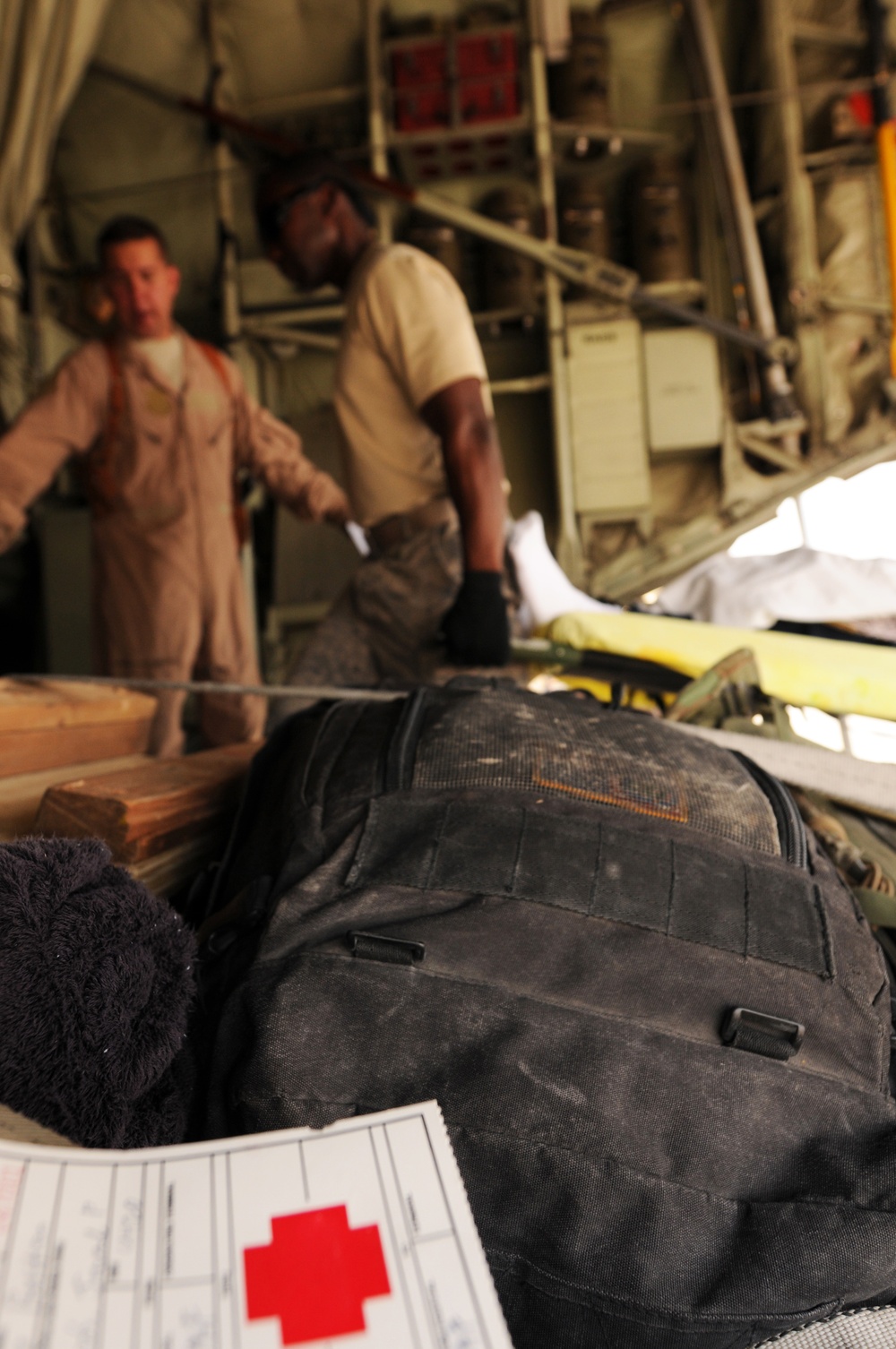 The image appears to be taken inside a military aircraft, likely during a medical evacuation (medevac) operation. In the foreground, a medical form bearing a red cross symbol is visible alongside a black tactical backpack. Two personnel in military attire are visible in the background, with what appears to be a litter or stretcher with a patient.