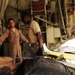 The image appears to be taken inside a military aircraft, likely during a medical evacuation (medevac) operation. In the foreground, a medical form bearing a red cross symbol is visible alongside a black tactical backpack. Two personnel in military attire are visible in the background, with what appears to be a litter or stretcher with a patient.