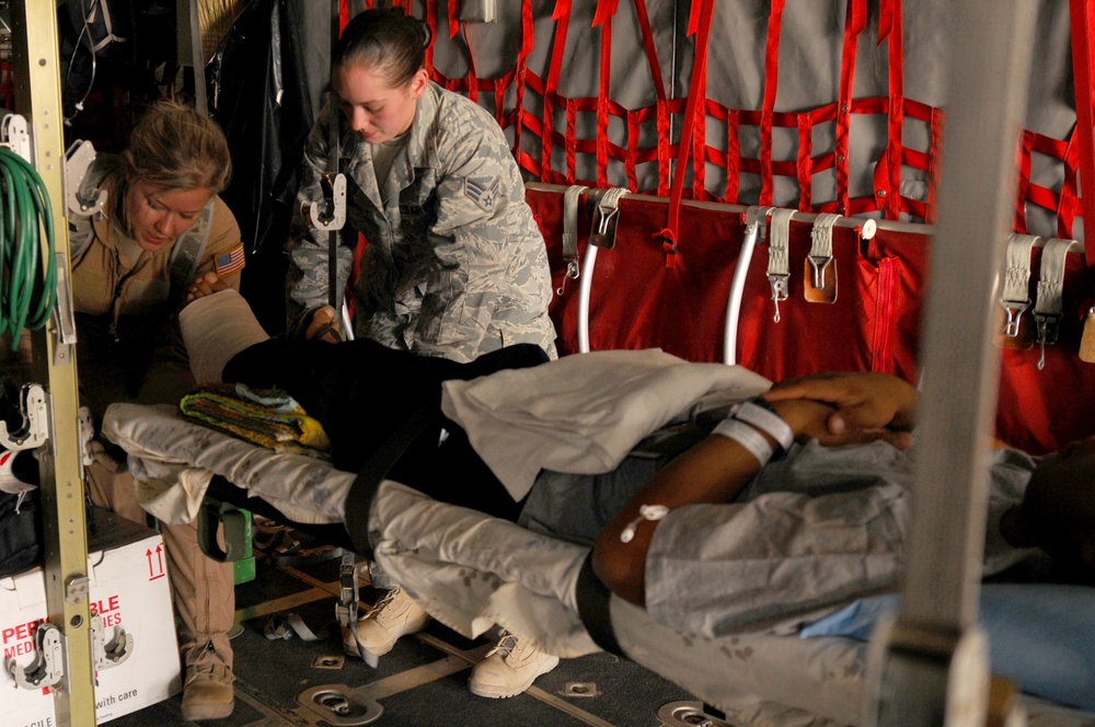 Two military medical personnel are attending to a patient on a litter inside what appears to be a military transport aircraft. One individual wears US Air Force ABU (Airman Battle Uniform) camouflage, while the other wears a tan/khaki uniform with a US flag patch. The patient is lying on a stretcher and appears to be receiving medical care during an aeromedical evacuation.