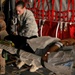 Two military medical personnel are attending to a patient on a litter inside what appears to be a military transport aircraft. One individual wears US Air Force ABU (Airman Battle Uniform) camouflage, while the other wears a tan/khaki uniform with a US flag patch. The patient is lying on a stretcher and appears to be receiving medical care during an aeromedical evacuation.