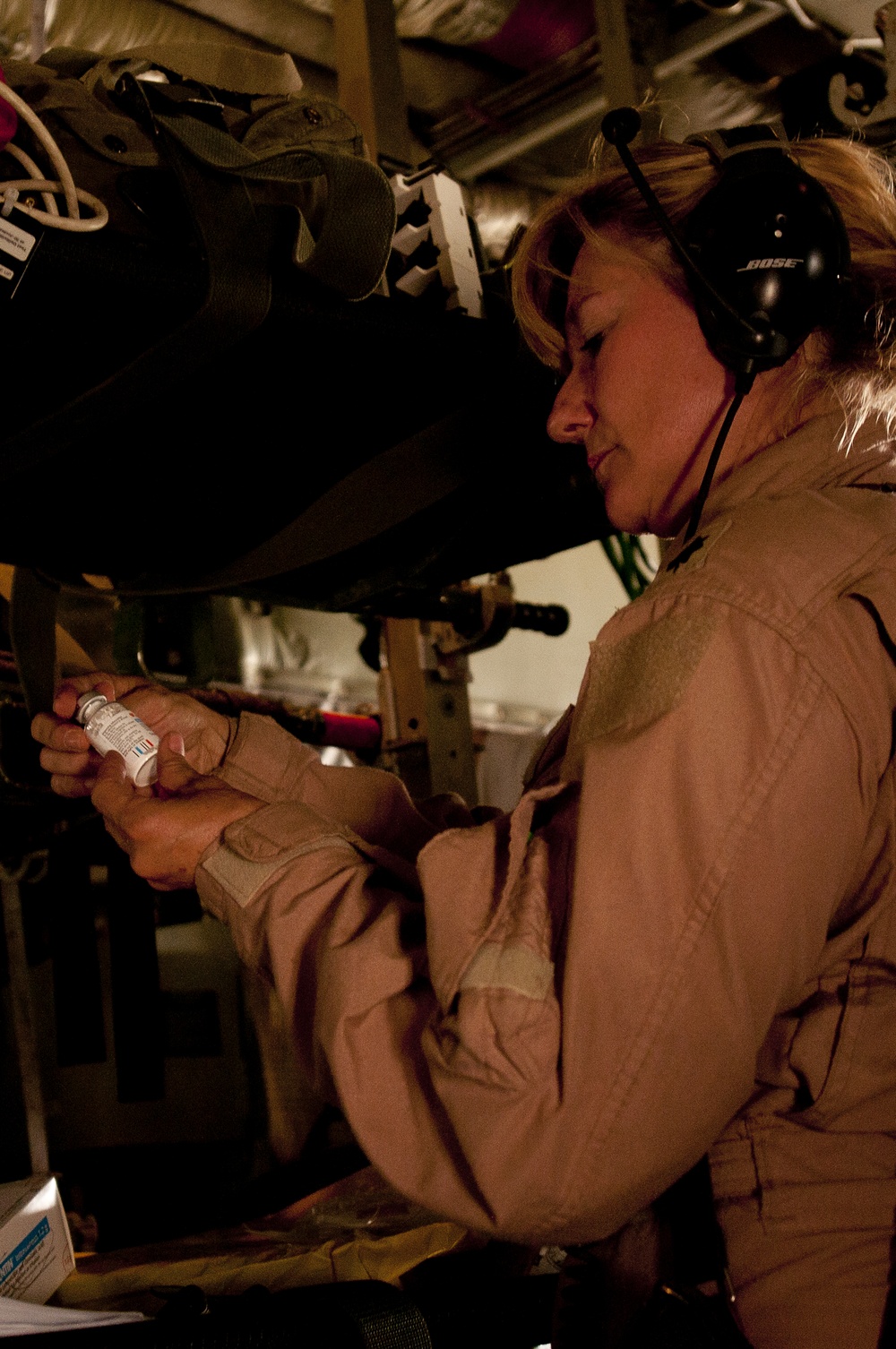 A woman wearing a tan/khaki flight suit and Bose aviation headset is examining a small medical vial inside what appears to be a military aircraft. She is focused on reading the label of the vial, suggesting a medical or aeromedical evacuation role. The interior environment shows military equipment, straps, and cargo fixtures consistent with a transport or medevac aircraft.