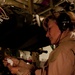 A woman wearing a tan/khaki flight suit and Bose aviation headset is examining a small medical vial inside what appears to be a military aircraft. She is focused on reading the label of the vial, suggesting a medical or aeromedical evacuation role. The interior environment shows military equipment, straps, and cargo fixtures consistent with a transport or medevac aircraft.