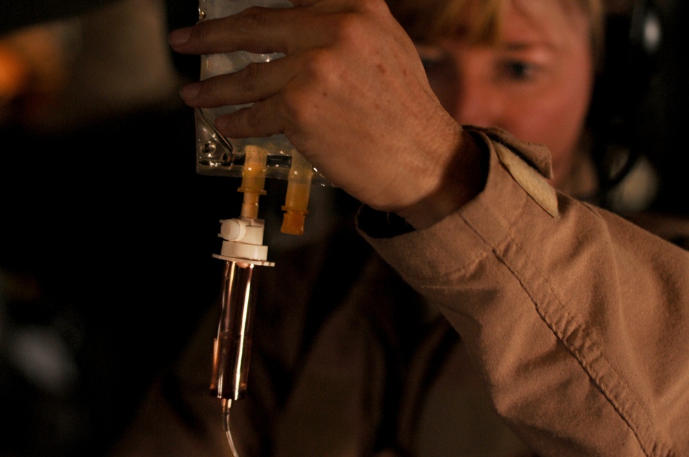 A person wearing a tan/khaki uniform is handling what appears to be an IV drip chamber or medical fluid administration set. The focus is on the medical equipment in the foreground, with the individual's face blurred in the background. The setting appears to be a low-light environment, possibly a medical or field setting.