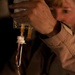 A person wearing a tan/khaki uniform is handling what appears to be an IV drip chamber or medical fluid administration set. The focus is on the medical equipment in the foreground, with the individual's face blurred in the background. The setting appears to be a low-light environment, possibly a medical or field setting.