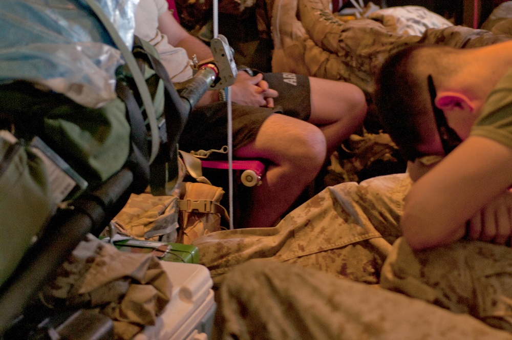 The image shows what appears to be a military medical evacuation or casualty care scenario in a low-light environment. Several individuals in military desert camouflage uniforms are present, with one person appearing to receive medical attention. The scene is crowded with military gear, backpacks, and equipment.