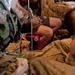 The image shows what appears to be a military medical evacuation or casualty care scenario in a low-light environment. Several individuals in military desert camouflage uniforms are present, with one person appearing to receive medical attention. The scene is crowded with military gear, backpacks, and equipment.