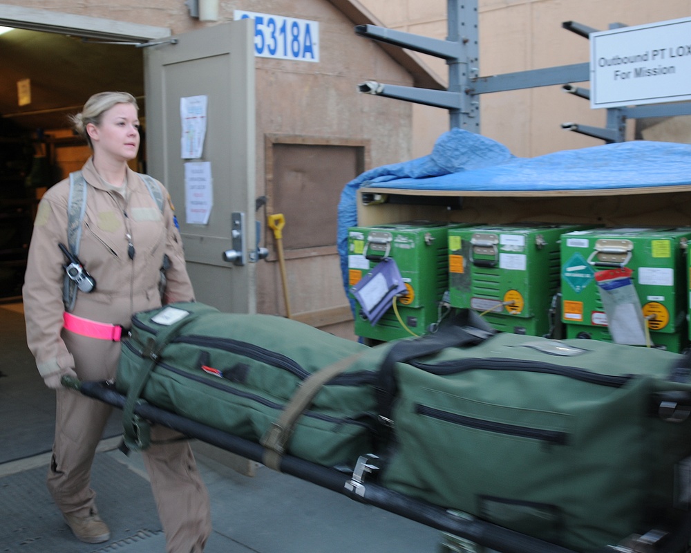 A female military or medical personnel member in tan/khaki flight suit is transporting equipment on a wheeled cart outside a building marked '5318A'. She is pulling a litter/stretcher loaded with olive drab duffel bags, with green medical or oxygen containers visible nearby on a separate cart. A sign reading 'Outbound PT LOX For Mission' is visible in the upper right, indicating an aeromedical evacuation or patient transport staging area.